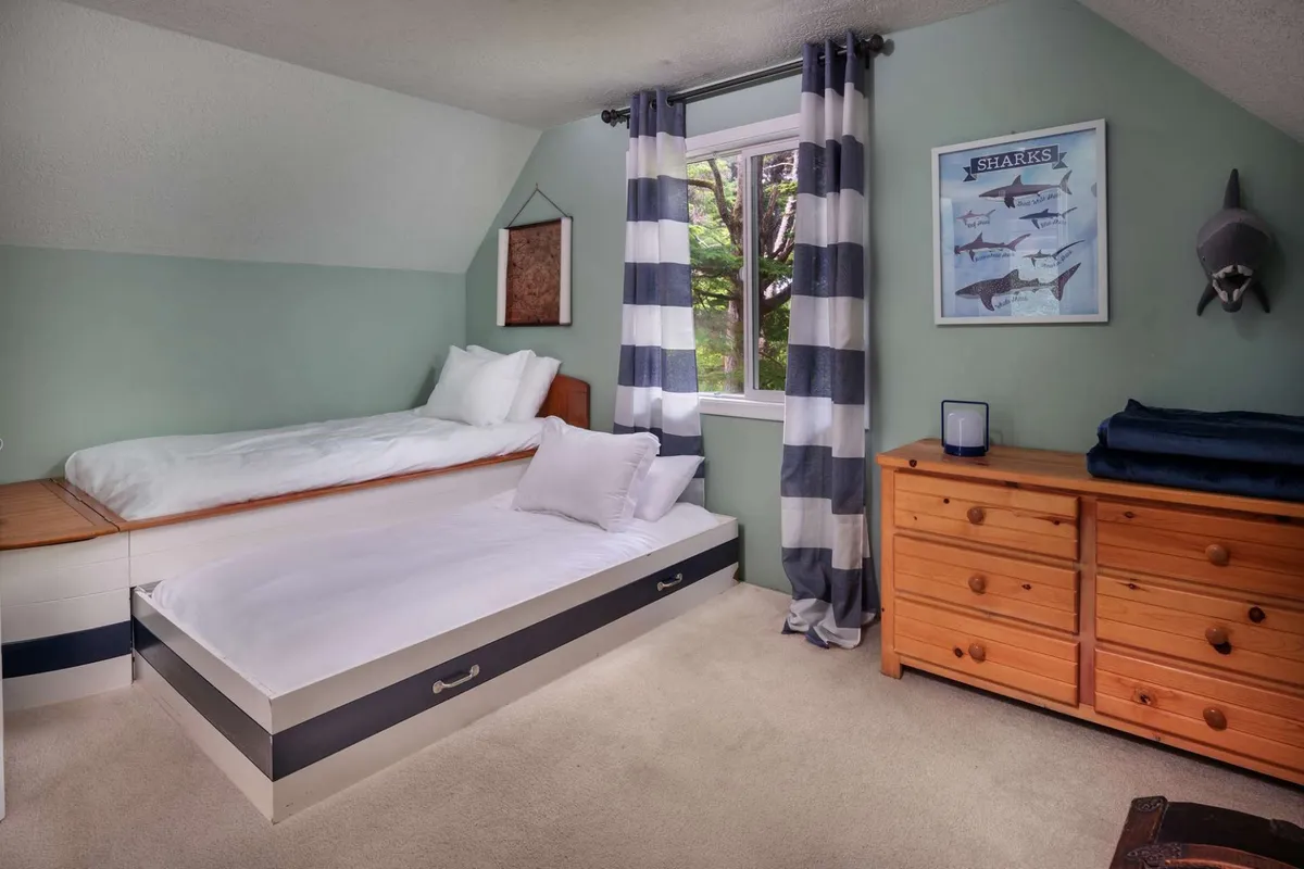 A cheerful bedroom with playful shark-themed wall art, a twin bed dressed in white linens, a window with striped curtains, and a wooden dresser. The space feels light, inviting, and ideal for children or friends sharing a room.