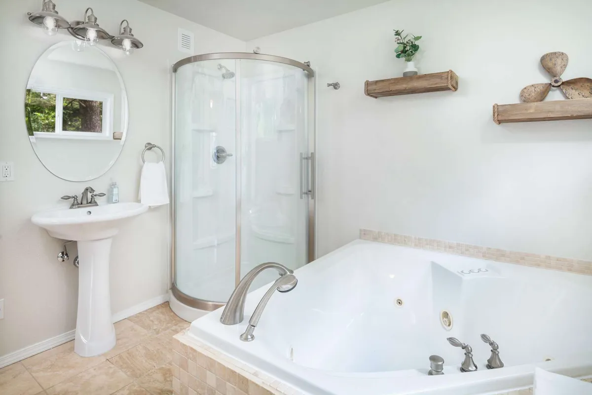 A tranquil, spa-like bathroom featuring a jetted Jacuzzi tub, a glass shower enclosure, a sink with a countertop, and wooden shelving. The serene design and thoughtful layout suggest a space for relaxation and rejuvenation after coastal adventures.