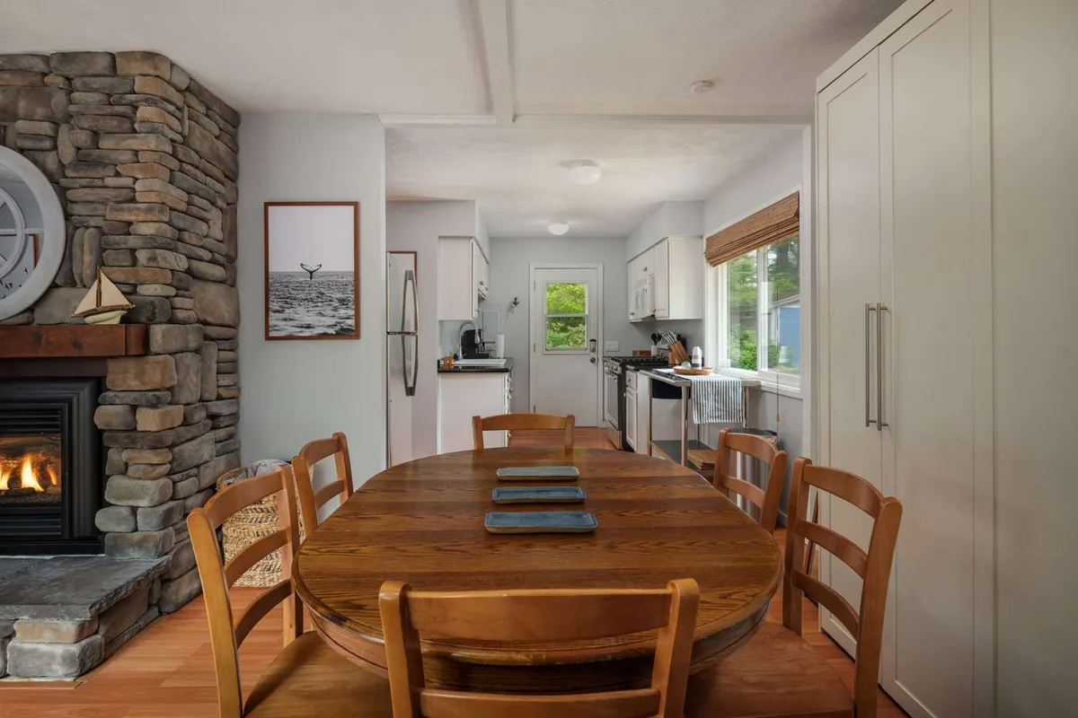 This image shows a welcoming dining area with a large wooden table set near a stone fireplace and open kitchen. Sunlight streams in, creating a warm, inviting ambiance perfect for gathering over meals or morning coffee.