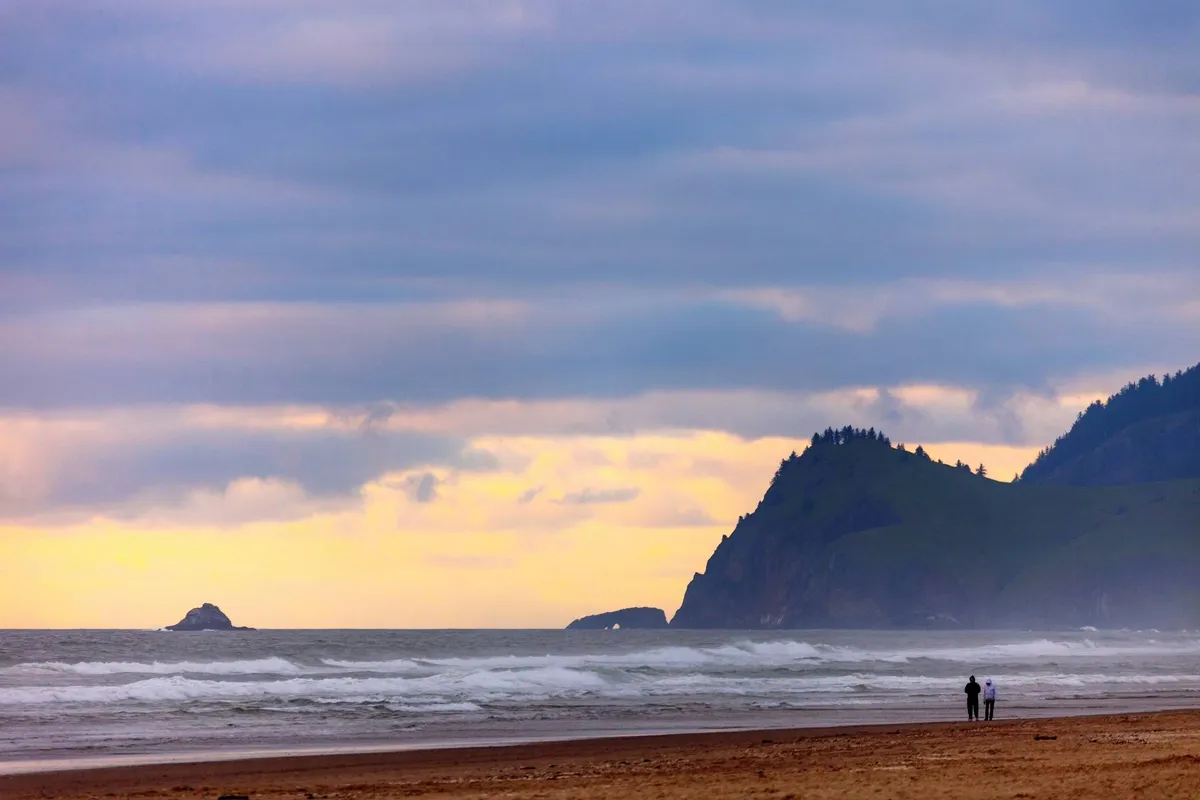 A breathtaking coastal view with ocean waves rolling up the sandy beach, distant cliffs, and a couple walking along the shoreline. The scene is awash in warm sunset light, evoking tranquility and the allure of beachside living.