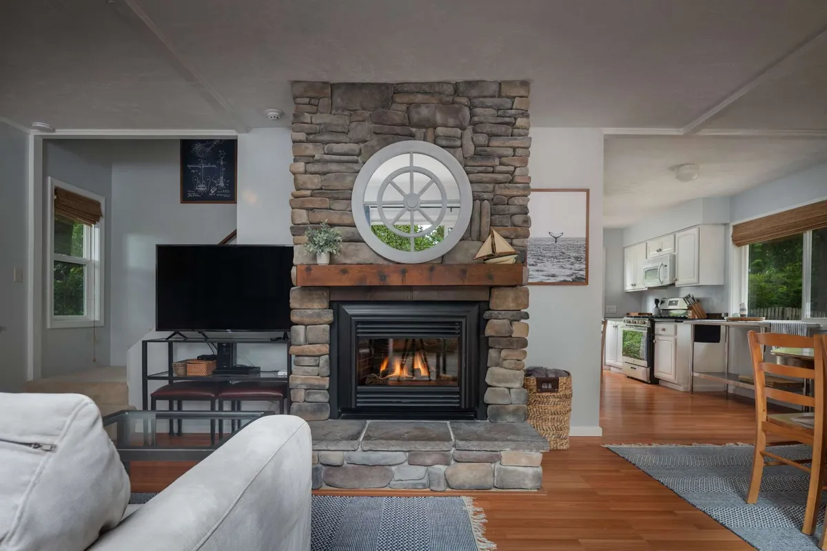 A bright, open-concept living area featuring a comfortable seating arrangement surrounding a fireplace and large flat-screen TV. In the background, the kitchen is visible, allowing for an integrated space where dining, relaxing, and socializing naturally flow together. Coastal touches in the decor and ample natural light create a welcoming environment.