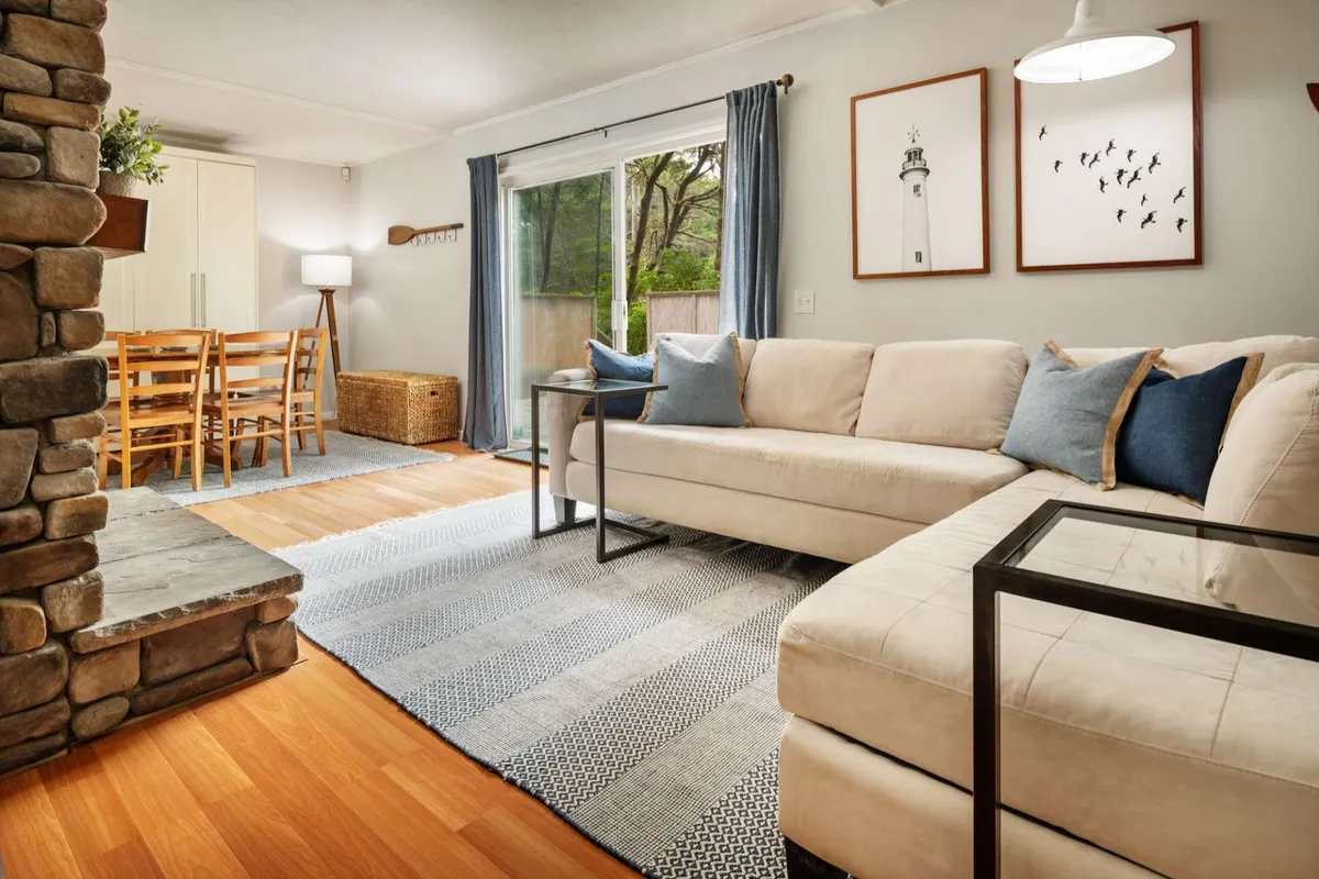 A stylish living room with plush sofas, a dining table for shared meals or games, and a stone fireplace. Large windows or sliding doors allow plenty of natural light, and tasteful coastal wall art enhances the seaside ambiance. The arrangement supports both intimate conversations and group activities.