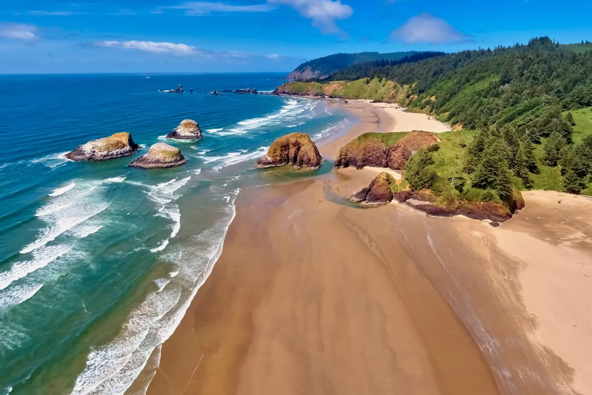 This image captures a serene stretch of sandy beach bordered by dramatic rock formations, gentle ocean waves, and a backdrop of lush forest. The scene conveys a sense of peacefulness, natural beauty, and the inviting shoreline just steps from the cottage.