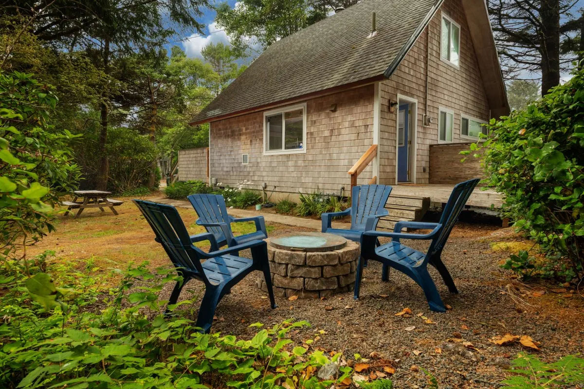 An inviting outdoor scene featuring blue Adirondack chairs arranged around a classic stone fire pit in the private backyard. The wooden cabin sits just beyond, while the lush greenery creates a peaceful, secluded atmosphere ideal for gatherings or quiet relaxation.