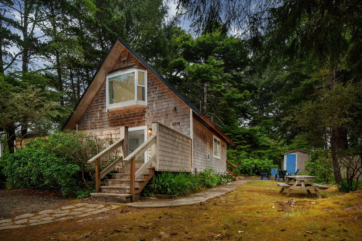 The hero image features a charming A-frame cottage set amidst lush greenery, with inviting wooden steps leading to the entrance. A picnic table and chairs are arranged for outdoor dining, and a cozy blue shed adds character to the peaceful garden setting. The scene exudes a sense of tranquility and warmth, perfectly suited for a relaxing getaway.