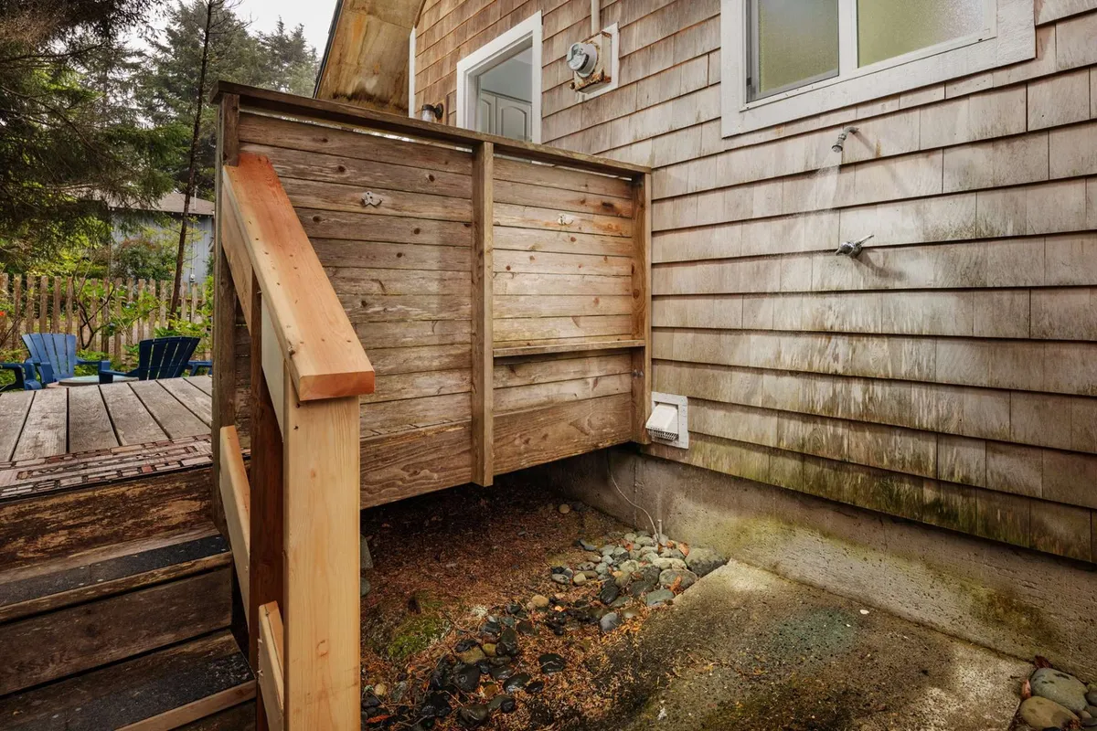 This exterior image captures the wooden steps leading up to a weathered siding and features a visible outdoor shower, perfectly positioned for rinsing off after time at the beach. The scene suggests a convenient spot for pets and people to clean up before heading indoors, with surrounding greenery adding a natural, relaxed vibe.
