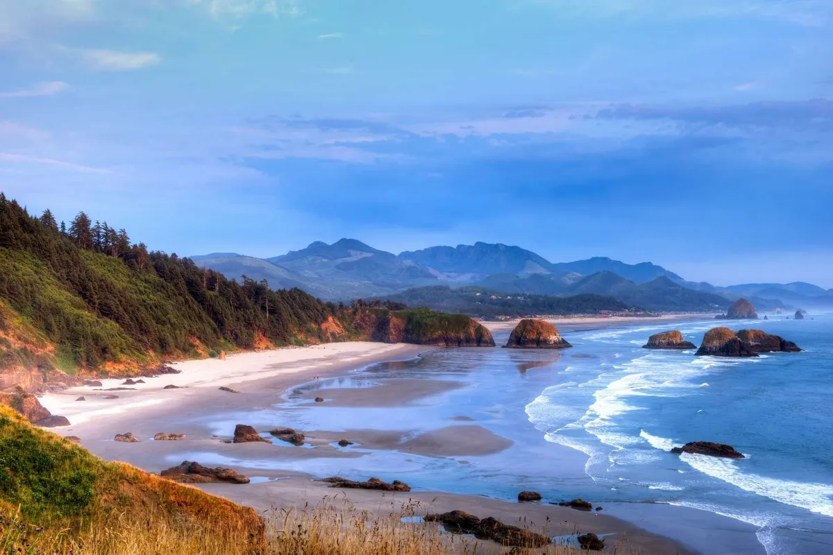 A sweeping coastal view showcasing distant green mountains, a rocky shoreline, expansive sandy beach, and rolling ocean waves. The tranquil mood is perfect for capturing the allure of sunset walks, beachcombing, and the natural drama of the Pacific coast.